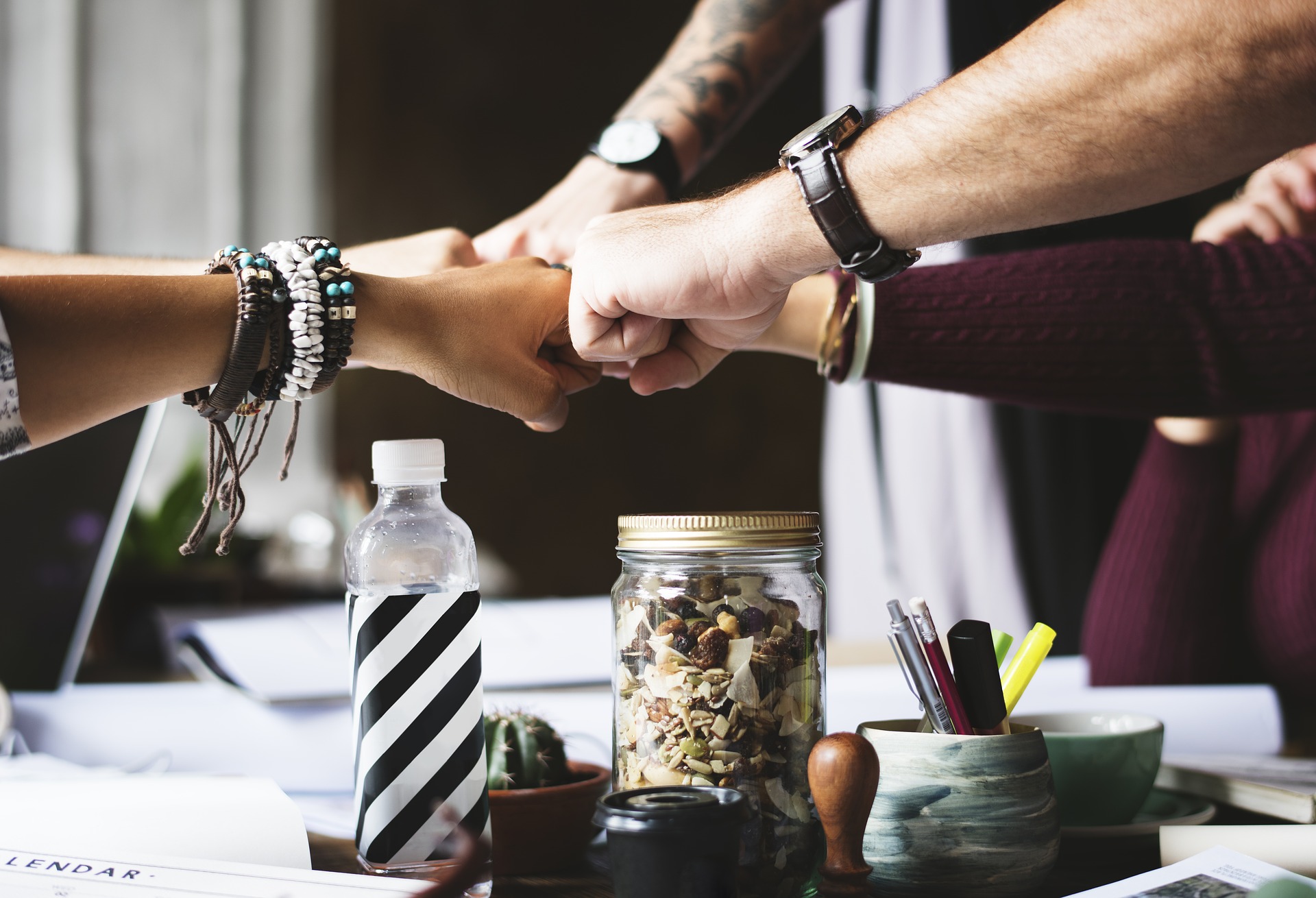 Team, Zusammenarbeit, Motivation, wir schaffen es, gemeinsam Arbeitstische mit Flasche, Stempel, Schreibutensilien, Kaktus. Fünf Menschen berühren mit jeweils einer Hand als Faust die anderen Fäuste