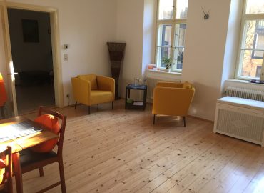 Besprechungsraum Ein Raum mit Parkettboden, weiße Wände, 2 gemütliche Stühle in gelb, Stehlampe, Beistelltisch, Tisch mit Stühlen